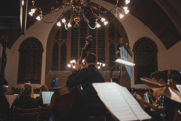 Een contrabassist speelt met zijn rug naar de camera in een sfeervol verlichte kerk, omringd door orkestleden en muziekstandaards onder een grote kroonluchter.