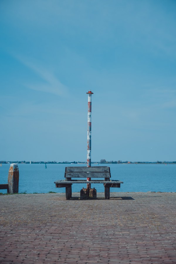 Een houten bank staat op een geplaveide kade bij een rood-witte meerpaal, met daarachter het rustige water en een strakblauwe lucht.