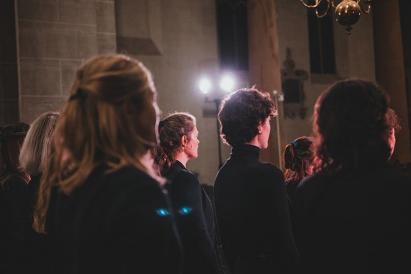 Koorleden in donkere kleding staan van achteren gezien in een schemerig gestemde kerkruimte, met warm spotlicht dat door de stenen pilaren schijnt.