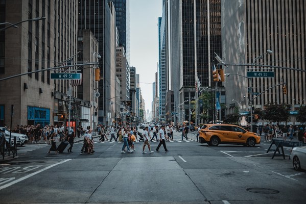 Een drukke straat in New York. We kijken de diepte in langs een aantal auto's en veel voetgangers