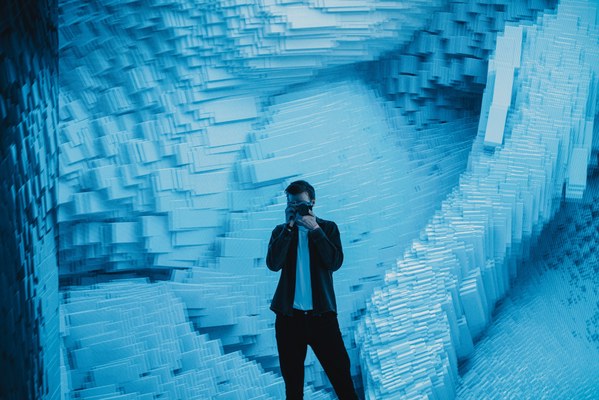 Een man in een zwart jasje maakt een foto met zijn camera, omringd door een golvende blauwe installatie van uitstekende blokjes.