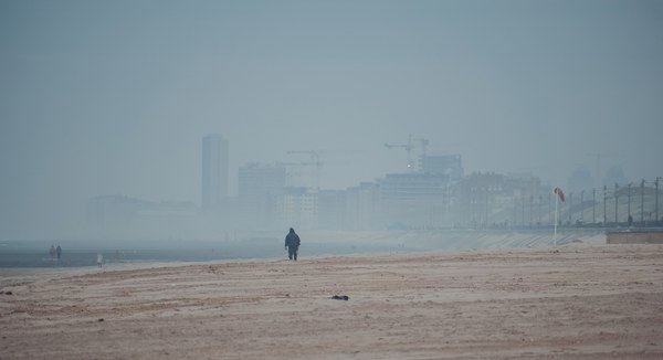 Een eenzame wandelaar loopt over een breed, mistig strand met op de achtergrond de vage skyline van de kuststad Middelkerke.