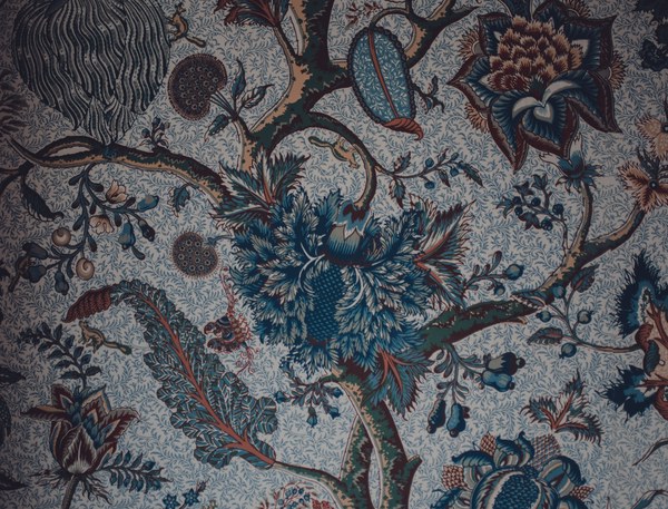 Rijk gedecoreerd textiel met een bloementak vol donkerblauwe en bruinrode bloemen, bladeren en knoppen tegen een fijn gebloemde lichte achtergrond.