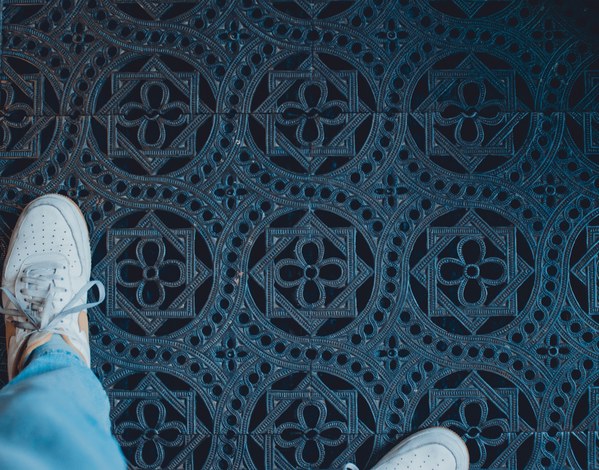 Vanuit vogelperspectief kijk je neer op witte sneakers en een blauwe spijkerbroek boven een sierlijke blauwe tegelvloer met klaverbladmotieven.