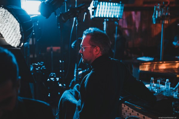 Man met bril en baard kijkt geconcentreerd opzij tussen filmlampen en apparatuur op een donkere set, verlicht door koelblauw licht.