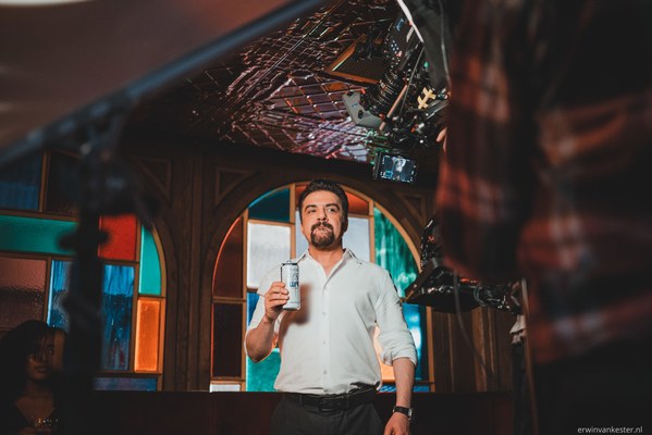 Man met baard in wit overhemd houdt tijdens een commercialopname een blikje omhoog, terwijl filmapparatuur en crew hem omringen.