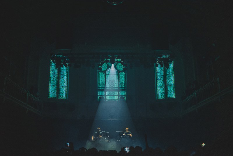Een foto van achterin de zaal van Paradiso. Een spot richt van bovenin de zaal op het podium. We zien de kerkramen van Paradiso.