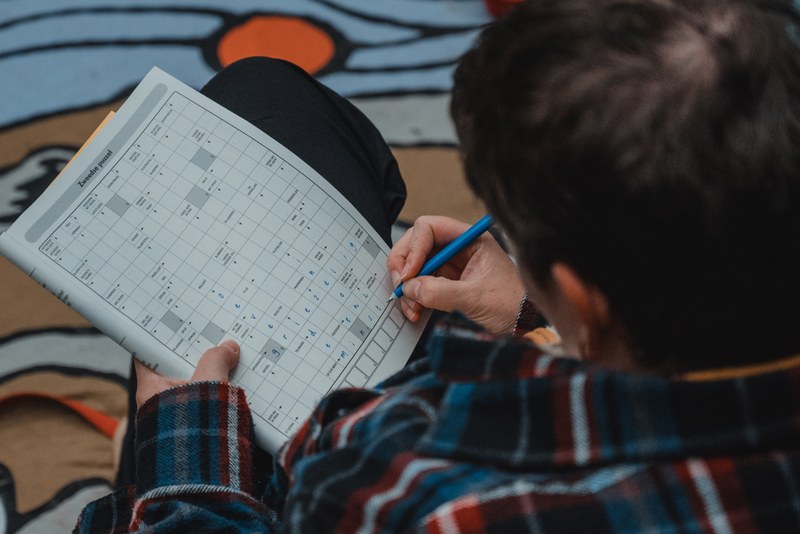 Van bovenaf gezien vult Veerle in een geruit flanellen hemd met een blauwe pen een puzzel in op een kleurrijk kleed.