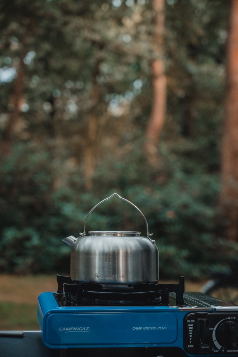 Een roestvrijstalen fluitketel staat op een blauw Campingaz-gasstel met een wazig bos van hoge dennenbomen op de achtergrond.