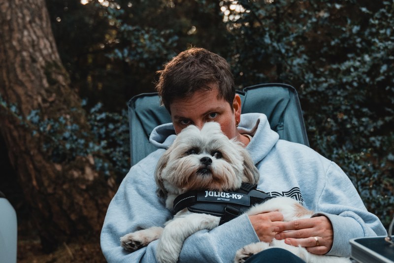 Een vrouw (Veerle) in een lichtblauwe hoodie zit in een campingstoel en houdt een hond (Kip) met Julius-K9-tuigje beschermend tegen zich aan tussen de bomen.
