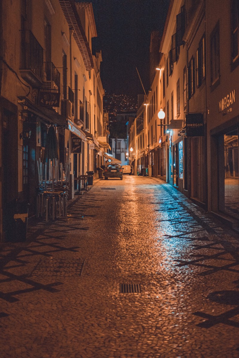 Een verlaten geplaveide straat in Funchal glinstert 's nachts onder oranje straatlantaarns, geflankeerd door winkels en een geparkeerde auto in de verte.