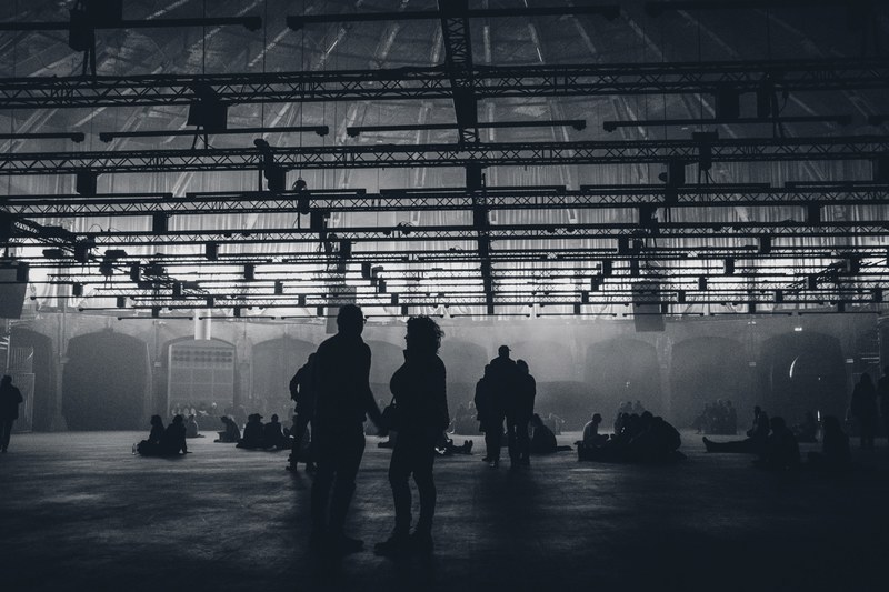 Silhouetten van mensen bewegen door een grote, industriële hal met een stalen dakconstructie, badend in diffuus licht en een mistige sfeer.