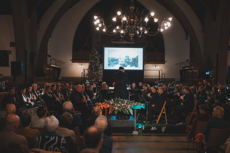 Een dirigent leidt een orkest tijdens een kerstconcert in een sfeervol verlichte kerk, met een kerstboom, projectiescherm en aandachtig publiek.