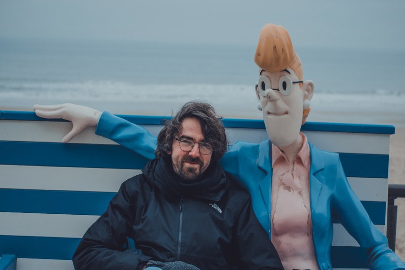 Een man met bril en donkere jas zit op een blauw-wit gestreepte bank naast een groot cartoonstandbeeld, met de mistige Noordzee op de achtergrond.