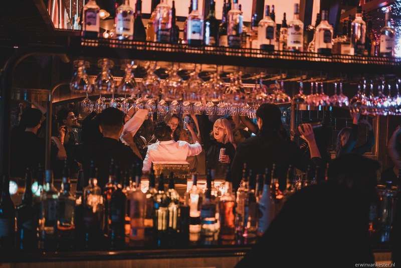 Bezoekers kletsen en drinken aan een sfeervolle bar met warme verlichting, omringd door talloze flessen en boven hen hangende wijnglazen.