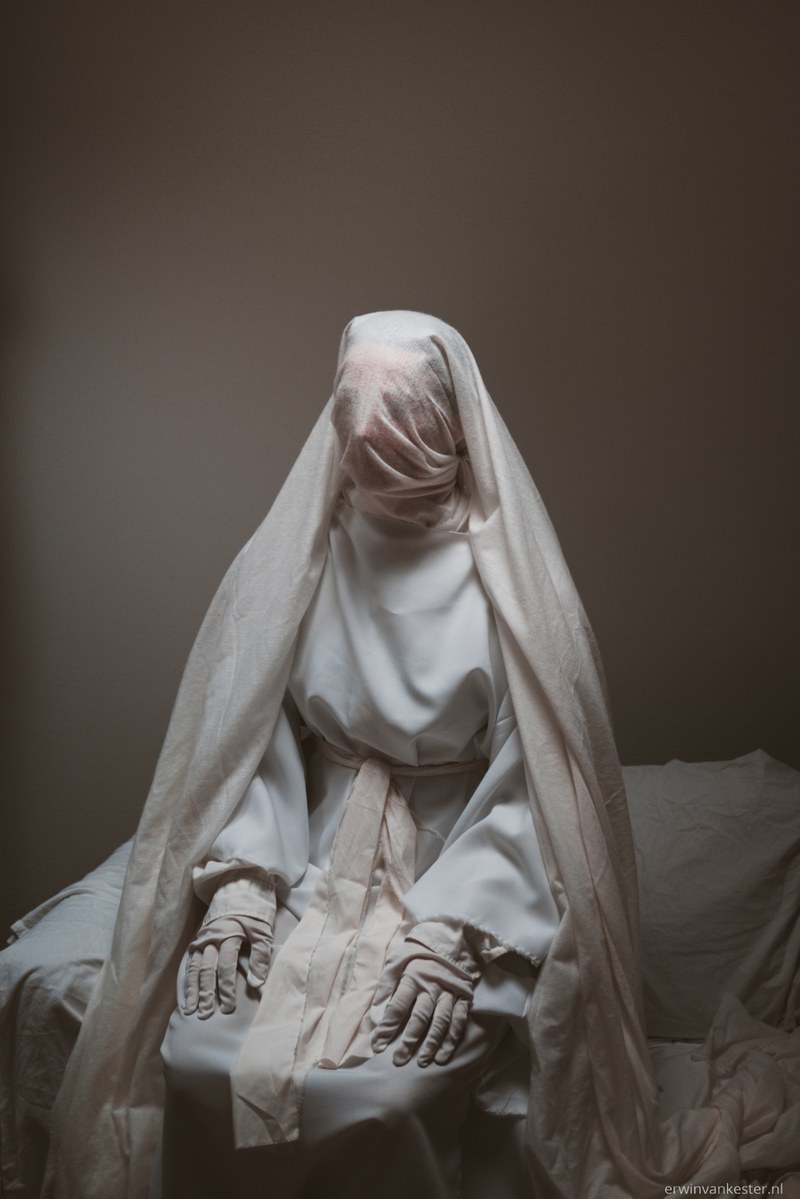 Een in witte doeken en een sluier gehulde figuur zit met de handen op de knieën op een bed, tegen een sobere bruine muur.