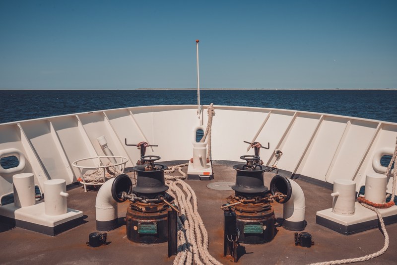 Het voordek van een schip met twee zwarte ankerlieren, opgerolde meertrossen en een witte reling, met daarachter een kalme blauwe zee onder een strakblauwe hemel.