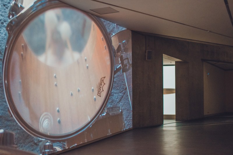 Een grote drum met Remo-vel domineert de voorgrond in een schemerige zaal met houten wanden en een verlichte doorgang op de achtergrond.