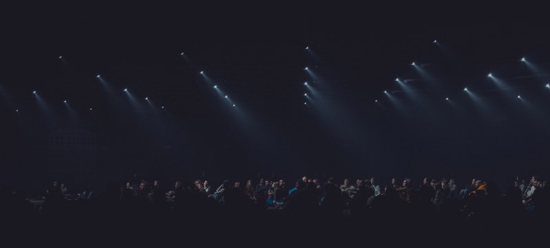 Rijen witte spotlights schijnen diagonaal door de duisternis boven een silhouet van een publiek tijdens de audiovisuele show Sora by Nonotak.