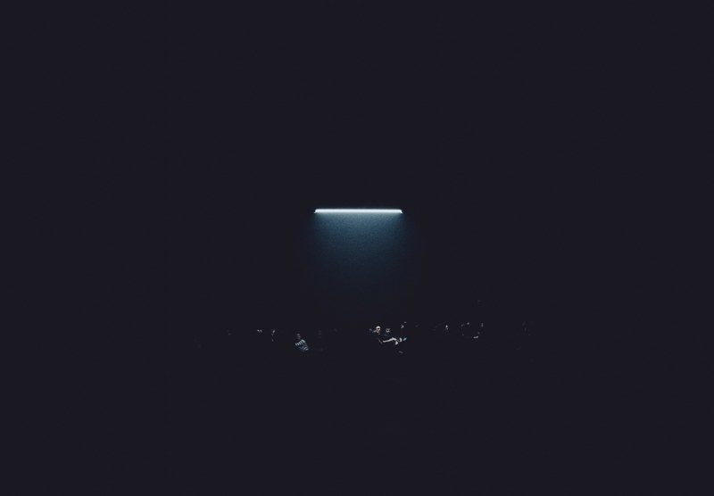 Een fel verlichte horizontale lichtbalk gloeit boven een donkere ruimte waarin de vage silhouetten van toeschouwers nauwelijks zichtbaar zijn.