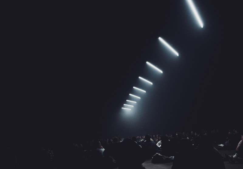 Een rij diagonaal opgehangen witte lichtstralen doorsnijdt de donkere ruimte boven de silhouetten van een publiek tijdens een audiovisuele voorstelling.