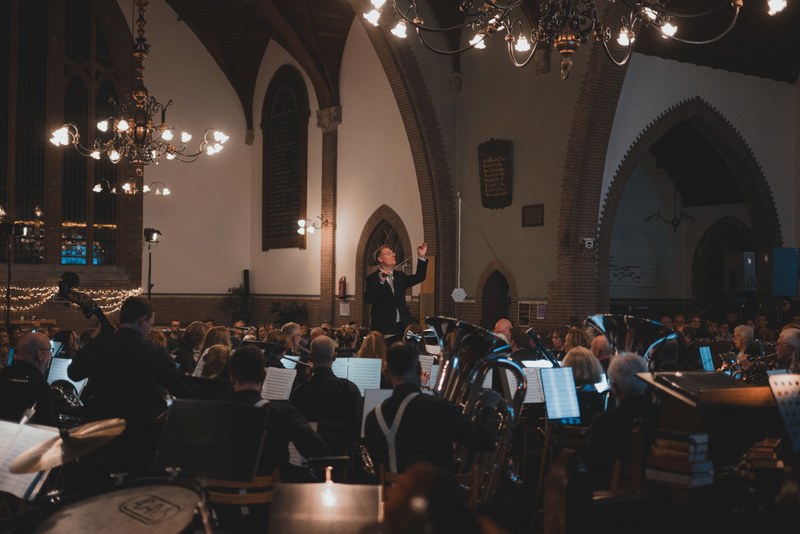 Een dirigent leidt met opgeheven armen een orkest tijdens een kerstconcert in een sfeervol verlichte kerk met kroonluchters en bogen.