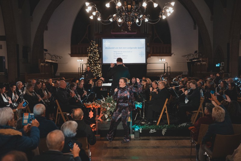 Een dirigent leidt een orkest in een sfeervol verlichte kerk tijdens een kerstconcert, terwijl het publiek toekijkt onder een grote kroonluchter.