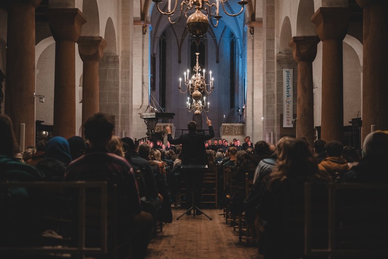 Een dirigent leidt vanaf een katheder een koor in een sfeervol verlichte kerk, terwijl het publiek in de banken toekijkt onder een gouden kroonluchter.