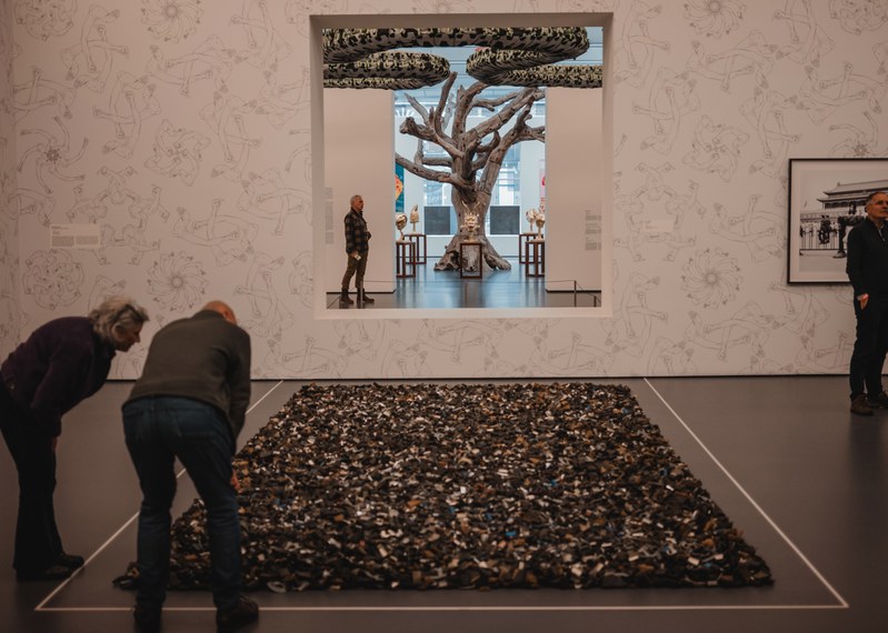Twee bezoekers buigen zich over een groot vloerwerk van talloze kleine objecten, terwijl door een muuropening een boomsculptuur en meer kunst zichtbaar zijn.