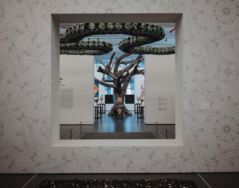 Door een vierkante muuropening met bloemenprint kijk je een museumzaal in, waar een grote boomsculptuur met groene bladerkronen centraal staat.