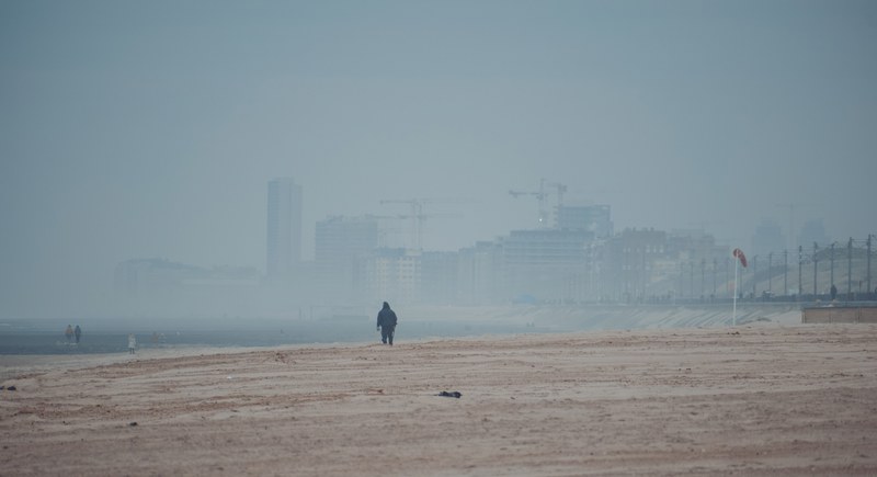 Een eenzame wandelaar loopt over een breed, mistig strand met op de achtergrond de vage skyline van de kuststad Middelkerke.