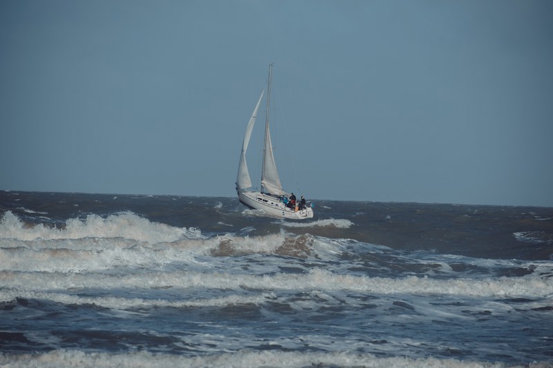 Een zeilboot met bemanning trotseert de ruwe, schuimende golven van de Noordzee onder een bewolkte grijsblauwe hemel bij Middelkerke.