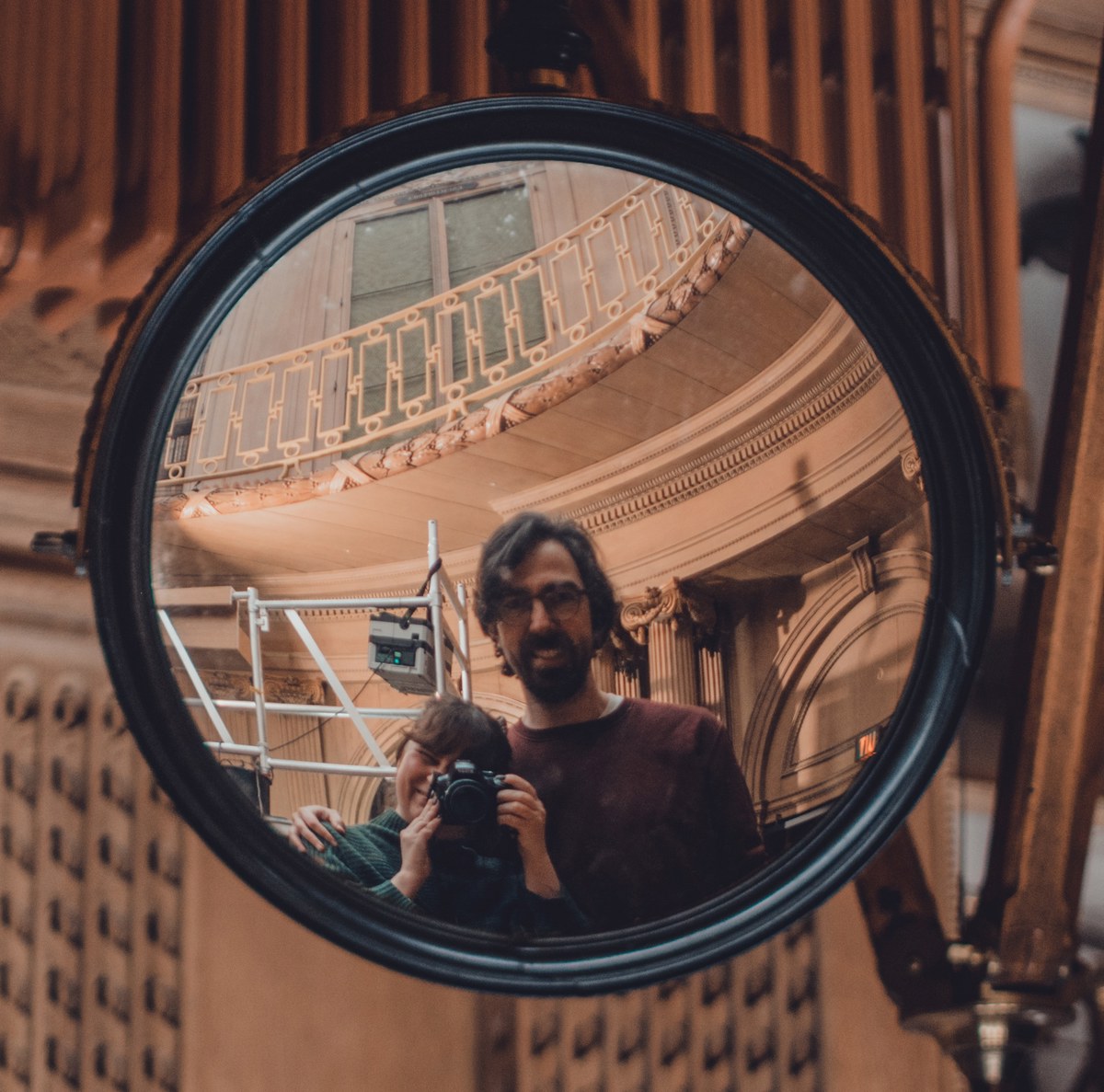Twee fotografen weerspiegeld in een grote ronde bolle spiegel, met een sierlijke balustrade en klassieke architectuur van het Teylers Museum op de achtergrond.