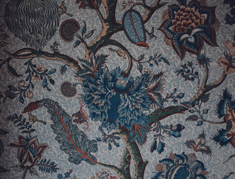 Rijk gedecoreerd textiel met een bloementak vol donkerblauwe en bruinrode bloemen, bladeren en knoppen tegen een fijn gebloemde lichte achtergrond.
