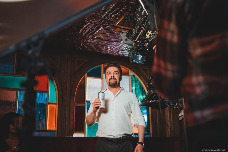 Man met baard in wit overhemd houdt tijdens een commercialopname een blikje omhoog, terwijl filmapparatuur en crew hem omringen.