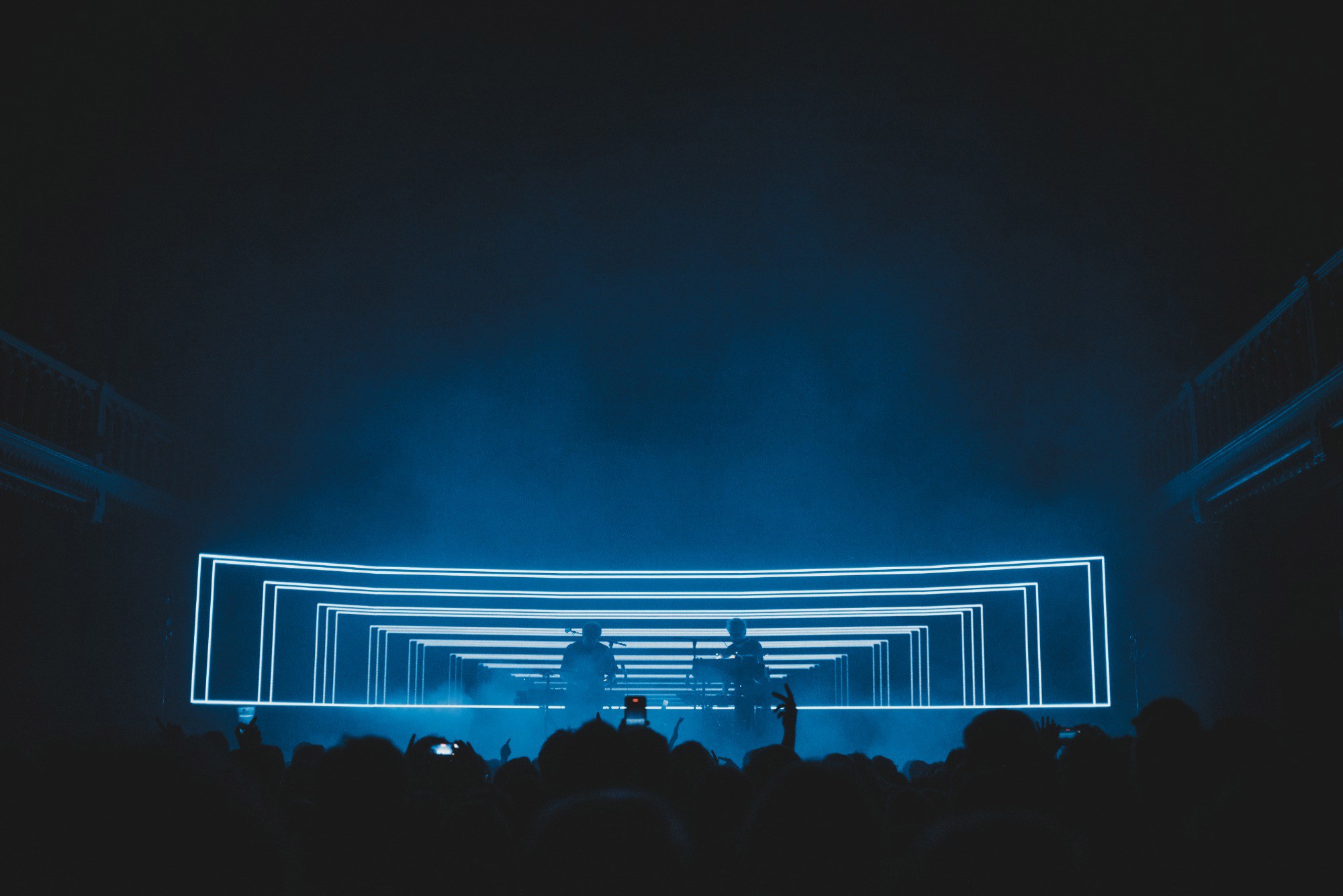 Een foto van achterin de zaal van Paradiso. We zien het publiek van achteren en de silhouetten van Weval. Ze worden van achter blauw belicht door een LED-scherm.